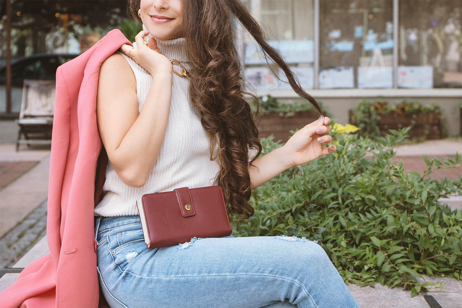 Protect Your Information This Holiday Season - A woman in a white top and jeans holds her pink blazer over her shoulder as she sits down on a stone wall. In her lap rests our Wine Madame Secretary Leather Clutch Wallet. 