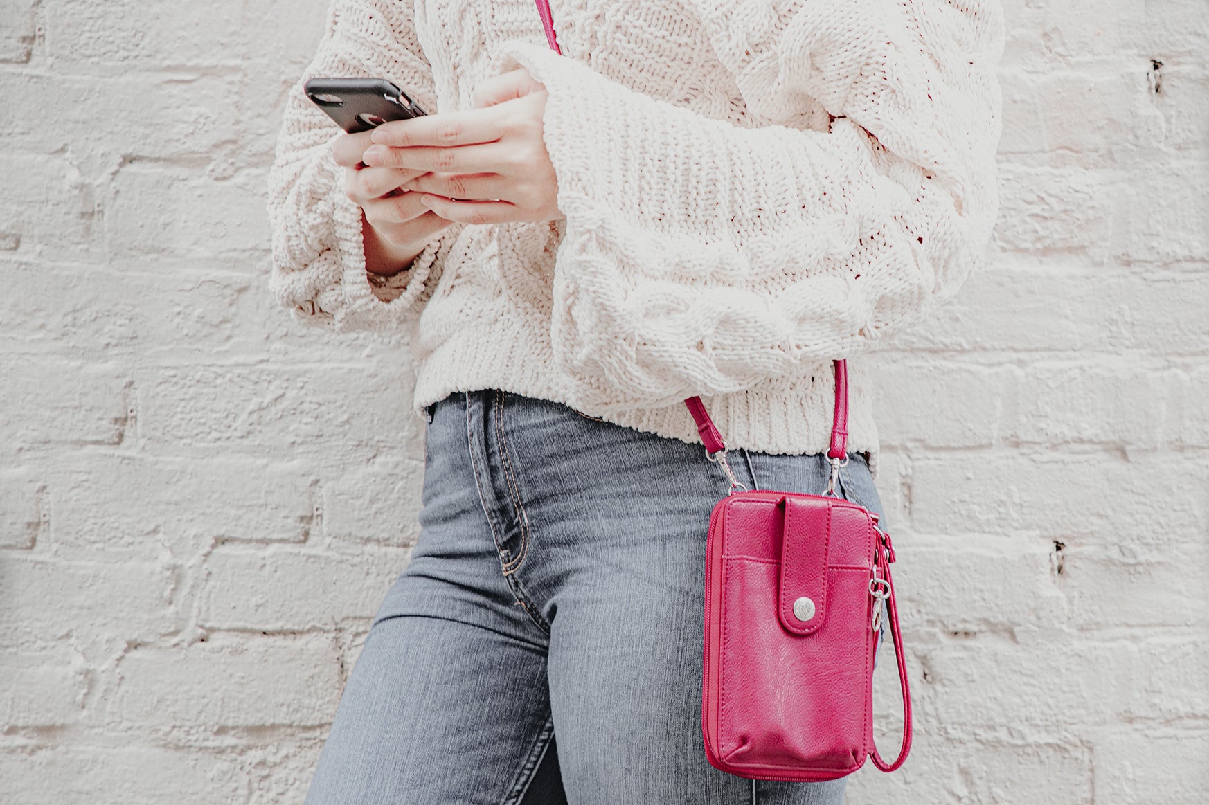 The Perfect Bags for Your Phone - A woman in a white sweater and jeans hangs her pink Jacqui Cell phone bag over her shoulder as she goes hands free and looks up something on her phone. 